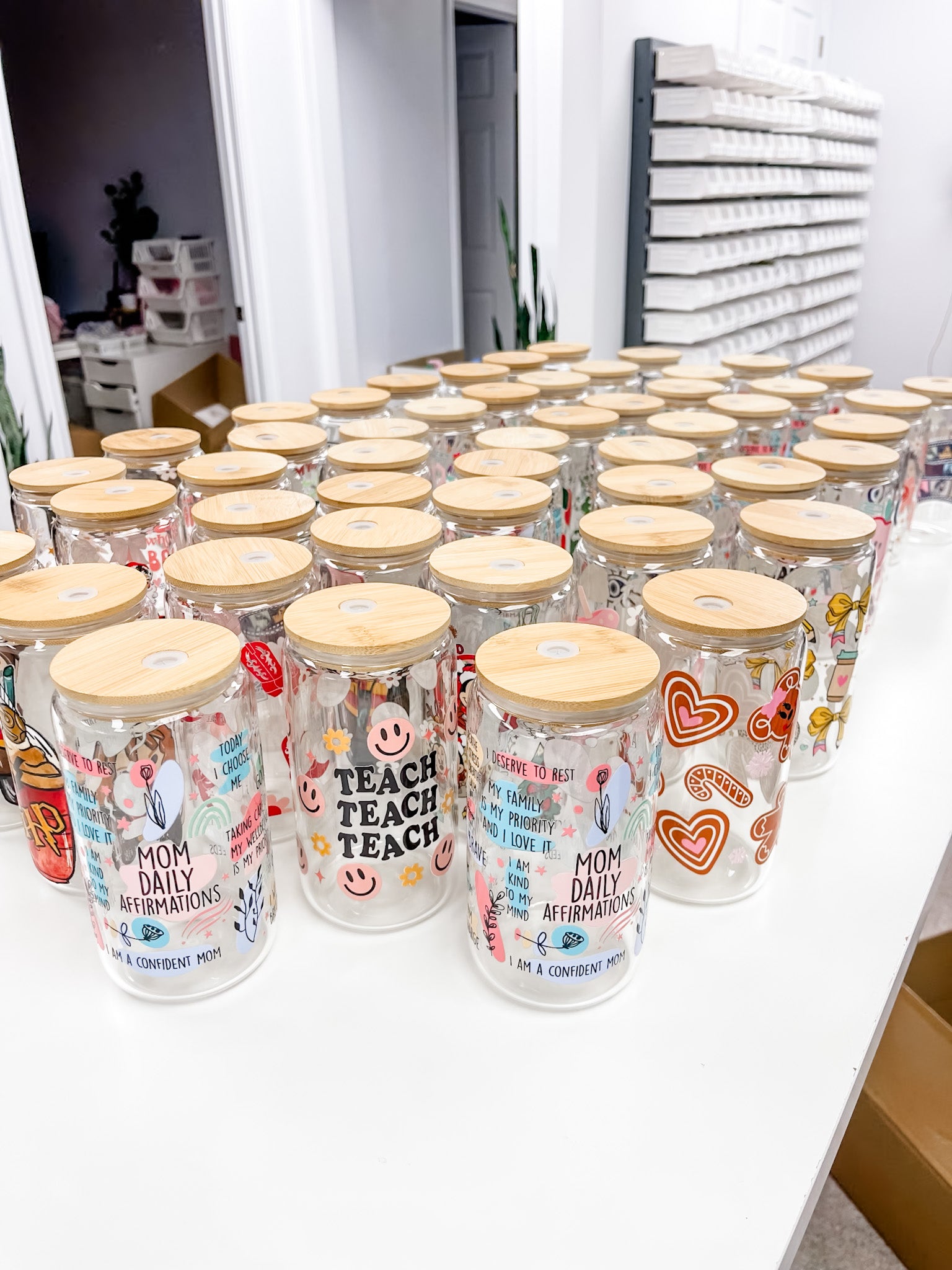 Multiple clear glass mason jars with light wood lids arranged on a white table in a retail or storage setting. The jars are decorated with colorful graphics, including hearts, smiley faces, and text such as &