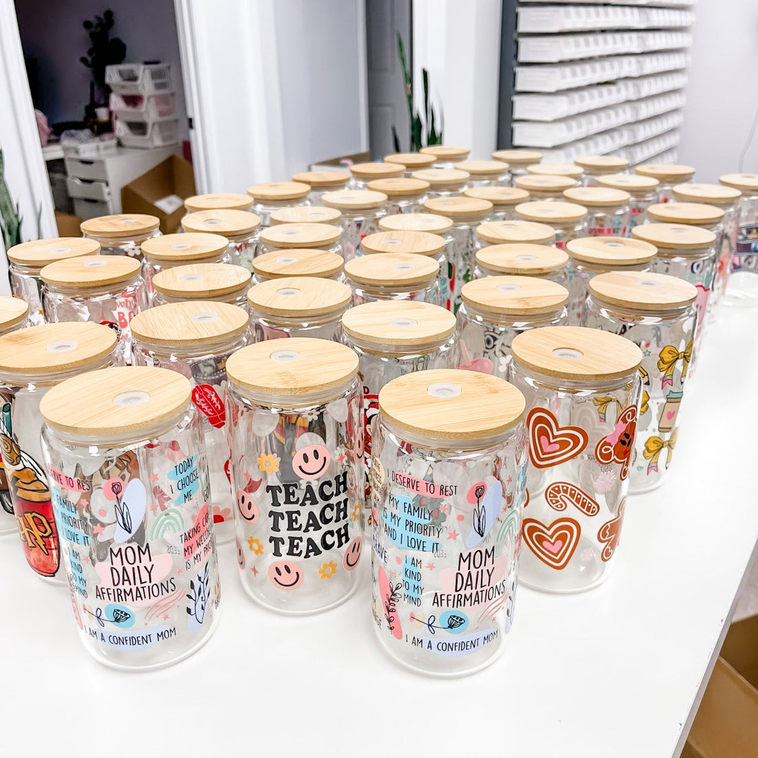 Multiple clear glass mason jars with light wood lids arranged on a white table in a retail or storage setting. The jars are decorated with colorful graphics, including hearts, smiley faces, and text such as &