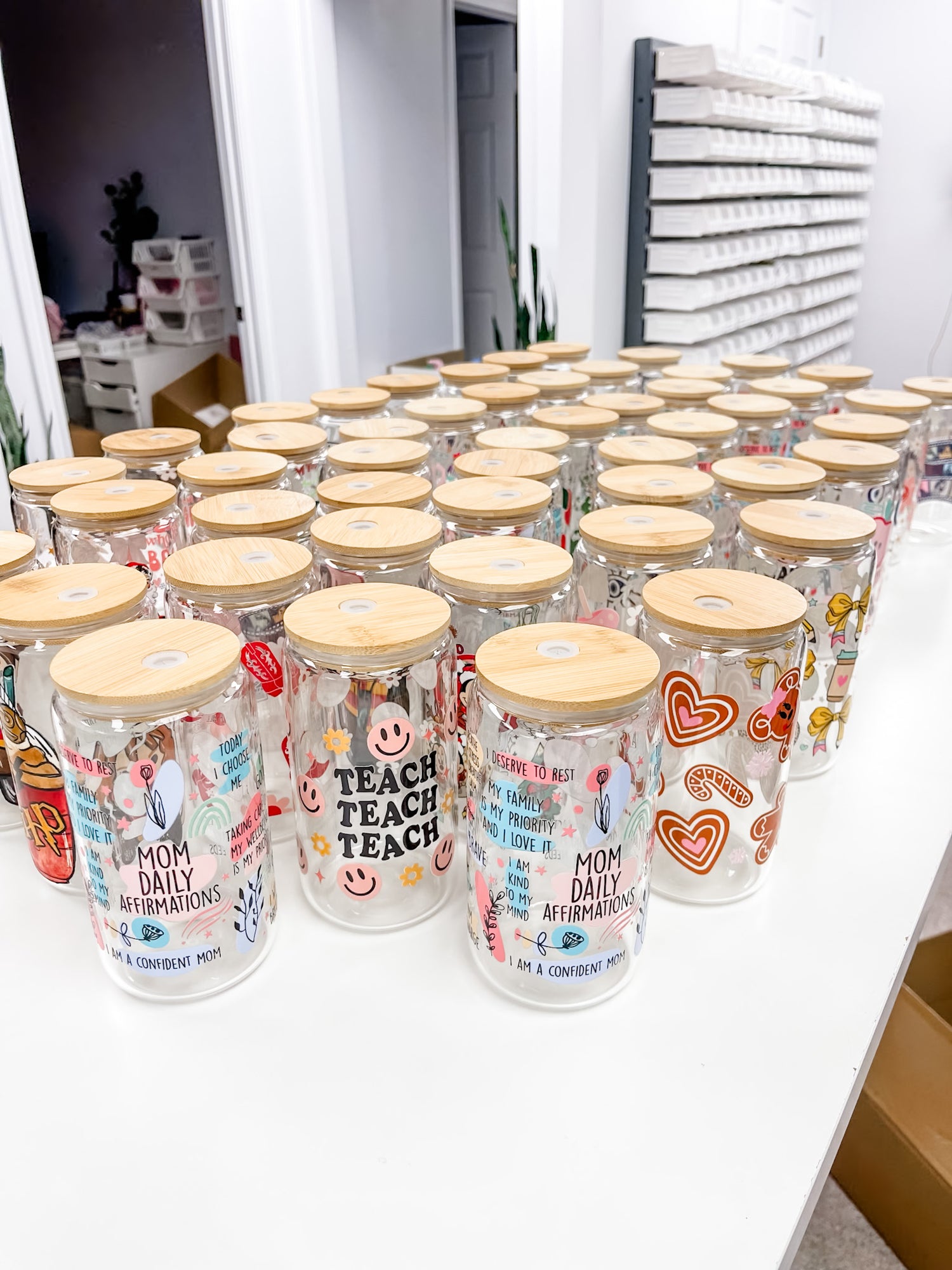 Multiple clear glass mason jars with light wood lids arranged on a white table in a retail or storage setting. The jars are decorated with colorful graphics, including hearts, smiley faces, and text such as &