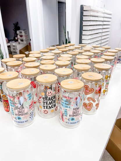 Multiple clear glass mason jars with light wood lids arranged on a white table in a retail or storage setting. The jars are decorated with colorful graphics, including hearts, smiley faces, and text such as &